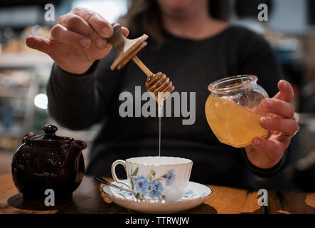 Mittelteil der Frau gießen Honig in Schale auf dem Tisch beim Sitzen im Cafe Stockfoto