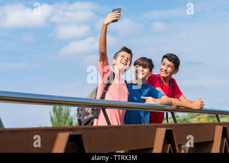 Der Schuljunge macht Selfie mit Freunden, während er an sonnigen Tagen auf der Fußgängerbrücke gegen den Himmel gelandet ist Stockfoto