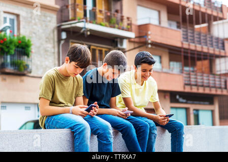 Freunde aus Smartphones beim Sitzen auf Stützmauer gegen Gebäude in der Stadt Stockfoto