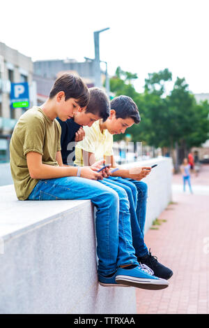 Freunde aus Smartphones beim Sitzen auf der Mauer gegen den klaren Himmel in der Stadt Stockfoto