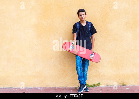 Portrait von Schüler holding Skateboard beim Stehen auf Fußweg gegen die Wand Stockfoto