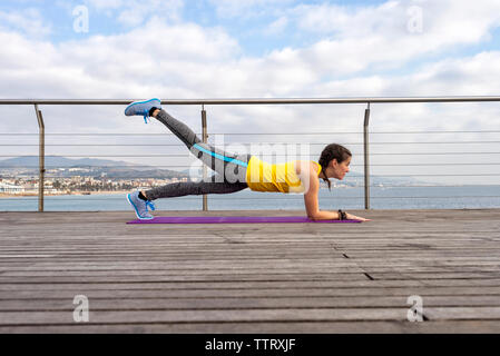 Seitenansicht der Frau in Sportswear sitzen auf Matten, Stretching auf Pier Stockfoto