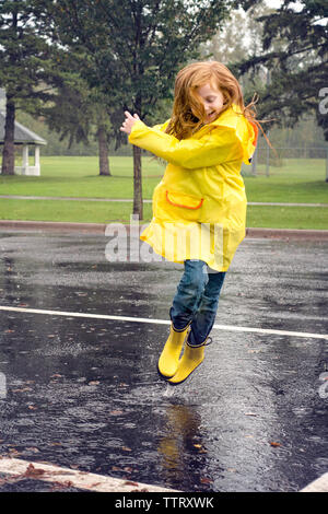 Fröhliches Mädchen Regenjacke tragen beim Springen auf der Straße bei Regen Stockfoto