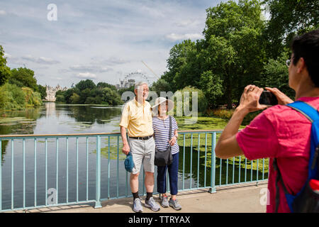 Sohn fotografieren Eltern mit Handy stehen auf eine Fußgängerbrücke über den Fluss gegen Himmel im Park während der sonnigen Tag Stockfoto