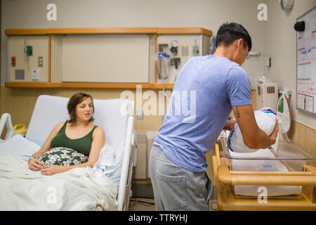 Ein Vater ruht sein neugeborenes in einem Krankenhaus Krippe, während seine Frau auf Blicken Stockfoto