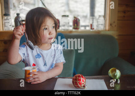 Ein Porträt von einem kleinen Mädchen Essen an einem Tisch vor dem großen Fenster Stockfoto