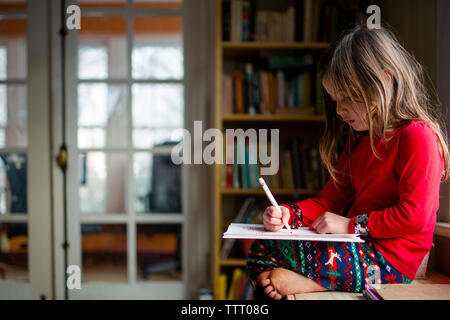 Ein kleines Mädchen sass barfuss durch ein Bücherregal schreiben in einem Notebook Stockfoto