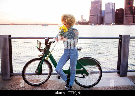 Riechende Frau Blumen beim Stehen mit dem Fahrrad gegen den Fluss Stockfoto