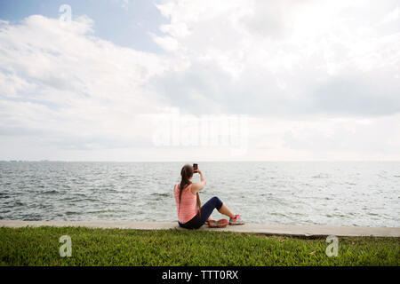 Ansicht der Rückseite Frau klicken selfie mit Smart Phone beim Sitzen am Ufer vor der See Stockfoto