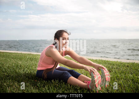 Junge Frau Zehen berühren beim Sitzen auf der Wiese vor der See Stockfoto