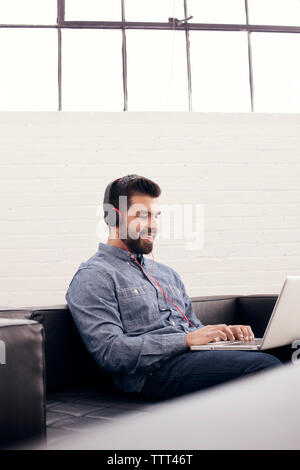 Lächelnder Mann mit Laptop das Tragen von Kopfhörern während kreative Büro zu sitzen Stockfoto