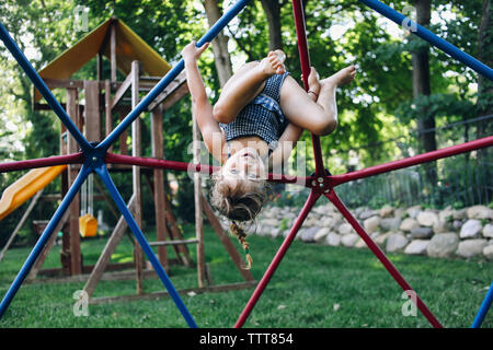 Portrait von Süß lächelnde Mädchen hing kopfüber an Jungle Gym gegen Bäume am Spielplatz Stockfoto