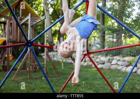 Portrait von niedlichen Happy girl hängend kopfüber auf Jungle Gym gegen Bäume am Spielplatz Stockfoto