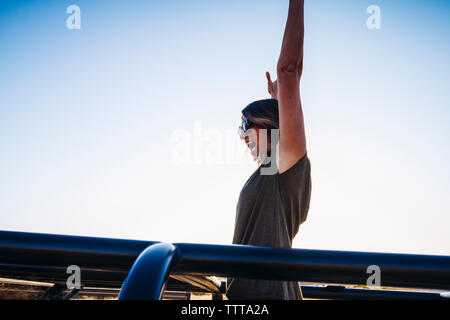 Low Angle View fröhliche Frau mit erhobenen Armen stehen in Geländewagen gegen den klaren Himmel Stockfoto