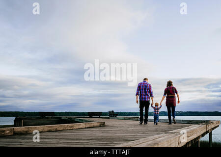 Ansicht der Rückseite des Familie Hand in Hand beim Gehen auf hölzernen Pier über den See gegen Sky Stockfoto