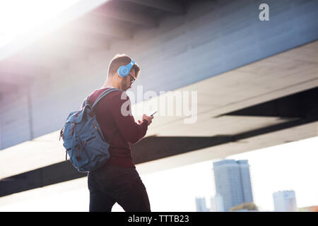 Junger Mann mit Phone beim Gehen gegen Brücke Stockfoto