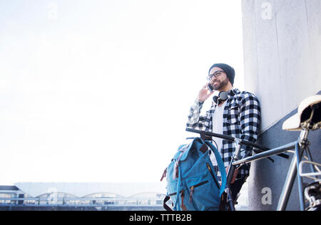 Junger Mann am Telefon sprechen und lehnte sich an der Wand mit dem Fahrrad gegen den klaren Himmel Stockfoto