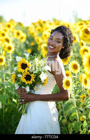 Portrait von lächelnden Braut holding Blumenstrauß im stehen inmitten von Sonnenblumen auf der Farm Stockfoto