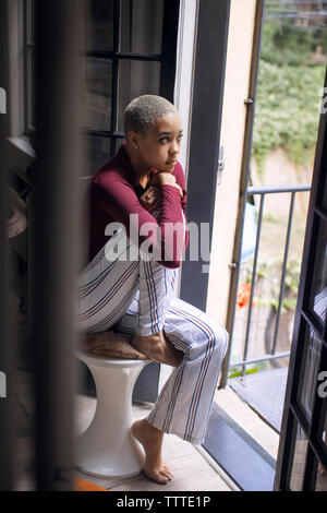 Nachdenkliche Frau weg, während auf Hocker zu Hause sitzen Stockfoto