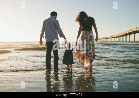 Rückansicht der Eltern halten sich an den Händen der Tochter beim Gehen im Meer bei Sonnenuntergang Stockfoto
