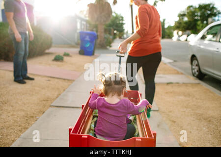 Verspielte Mutter ziehen Tochter im toy Zirkuswagen mit Vater stehen im Hintergrund Stockfoto