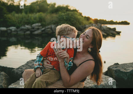 Glückliche Mutter mit Sohn sitzen auf den Felsen am Seeufer bei Sonnenuntergang Stockfoto