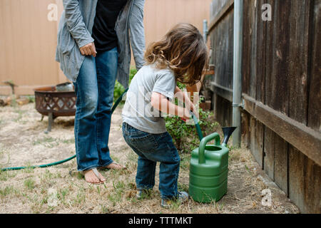 Die ganze Länge der Tochter mit Gartenschlauch Bewässerung zu füllen können, während man durch die Mutter im Hinterhof Stockfoto
