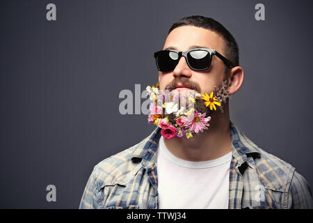 Schöner Mann in Sonnenbrille mit Bart von Blumen auf dunklem Hintergrund Stockfoto