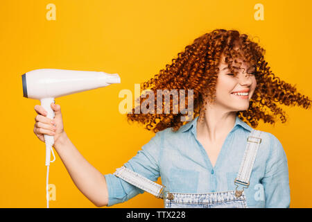 Curly rothaarige Frau mit Fön auf gelben Hintergrund. Die perfekte Locken Stockfoto