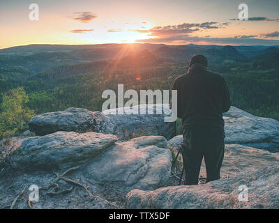 Fotograf mit Stativ auf einer Klippe und Denken. Verträumt neblige Landschaft orange misty Sonnenaufgang in einem schönen Tal, unten Stockfoto