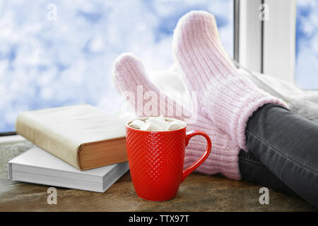 Menschliche Beine in gestrickten Socken auf der Fensterbank neben Kaffee und Bücher Stockfoto