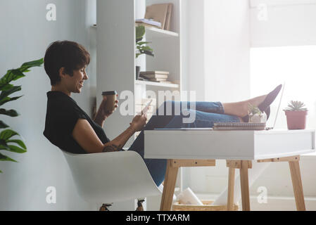 Hübsche Junge lächelnd Geschäftsfrau in einem Stuhl sitzend mit Ihrem Smartphone mit den Beinen über dem Schreibtisch in warme Hintergrundbeleuchtung Stockfoto