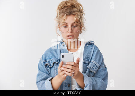 Portrait besorgt Intensive gut aussehende Stilvolles, modernes 20s junge Frau blonde lockige unordentlich bun Brille Jeansjacke Blick gestört nervös Smartphone Stockfoto