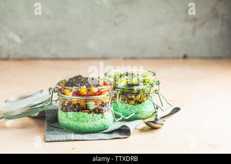 Zwei Chia Samen Pudding mit Matcha Grüntee, Kiwi und Müsli, orange in Gläsern auf Hellrosa konkreten Hintergrund. Gesundes Frühstück. Stockfoto