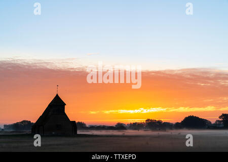 Der hl. Thomas Becket Kirche auf Romney Marsh. Die Kirche auf dem Sumpf bei Sonnenaufgang. Sunup neben der Kirche mit Band der hellen orange Cloud. Nebel auf dem Boden Stockfoto