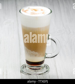 Schaumige, mehrschichtige Cappuccino in einem klaren Glas Becher mit Zimt bestreut oben auf weißem Holz- Hintergrund Stockfoto