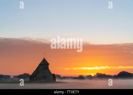 Der hl. Thomas Becket Kirche auf Romney Marsh. Die Kirche auf dem Sumpf bei Sonnenaufgang. Sunup neben der Kirche mit Band der hellen orange Cloud. Nebel auf dem Boden Stockfoto