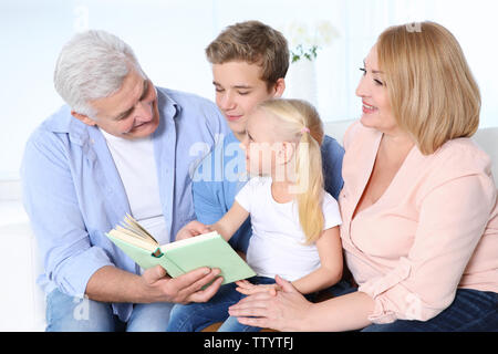 Niedliche Kinder mit Großeltern lesen Buch beim Sitzen auf einem Sofa im Wohnzimmer. Stockfoto