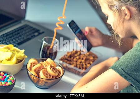 Mädchen arbeitet an einem Computer und frisst fast food. Ungesundes Essen: Chips, Kekse, Bonbons, Waffeln, Cola. Junk Food, Konzept. Stockfoto