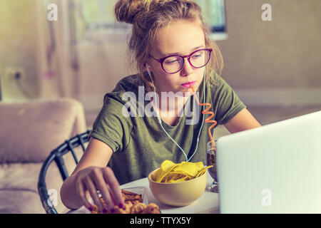 Mädchen arbeitet an einem Computer und frisst fast food. Ungesundes Essen: Chips, Kekse, Bonbons, Waffeln, Cola. Junk Food, Konzept. Stockfoto