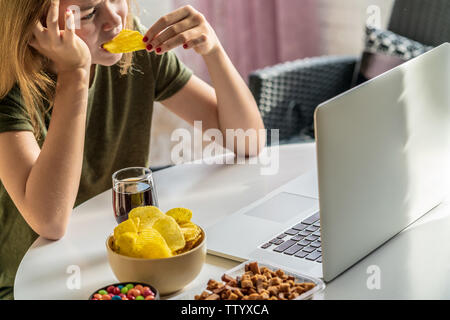 Mädchen arbeitet an einem Computer und frisst fast food. Ungesundes Essen: Chips, Kekse, Bonbons, Waffeln, Cola. Junk Food, Konzept. Stockfoto