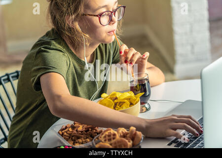 Mädchen arbeitet an einem Computer und frisst fast food. Ungesundes Essen: Chips, Kekse, Bonbons, Waffeln, Cola. Junk Food, Konzept. Stockfoto