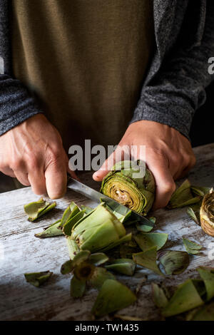 Nahaufnahme von einem kaukasischen Mann Schneiden mit einem Messer einige Rohe frische Artischocken, auf einem weißen rustikalen Holztisch Stockfoto