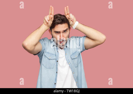 Portrait von Angry gut aussehender bärtiger junger Mann in blau casual Shirt stehend mit Hörnern Geste auf dem Kopf und Kamera mit ernsten Gesicht. Stockfoto