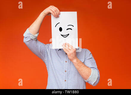 Jungen Kerl versteckt sein Gesicht unter gemalt winking Lächeln auf weißem Papier, orange Hintergrund, Panorama Stockfoto