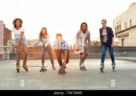 Sommer Urlaub, Vermietungen und Freundschaft Konzept. Gruppe von lächelnden Teenager mit Rollschuhen und Skateboard reiten im Freien im Sommer am Abend Stockfoto