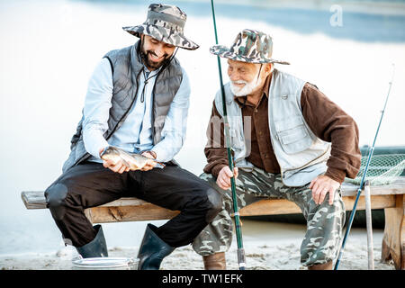 Gerne Großvater mit Sohn Holding frisch gefangenen Fisch, während sitzen zusammen in der Nähe des Sees in den frühen Morgenstunden Stockfoto