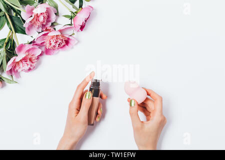 Weibliche Hände, die Make-up-blending schwamm Applikator und Gesichtsbehandlung Foundation Cream in der Nähe von Bouquet von rosa Pfingstrosen auf weißem Hintergrund, flach. Stockfoto