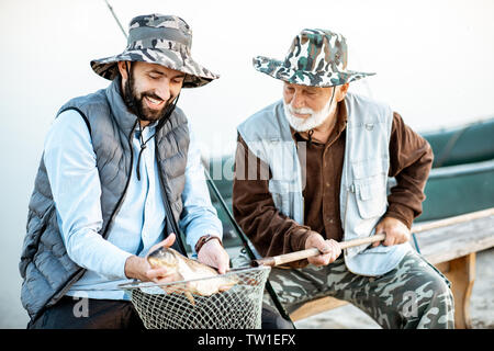 Gerne Großvater mit Sohn Holding frisch gefangenen Fisch, während sitzen zusammen in der Nähe des Sees in den frühen Morgenstunden Stockfoto