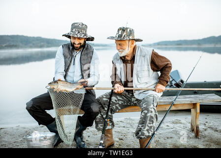 Gerne Großvater mit Sohn Holding frisch gefangenen Fisch, während sitzen zusammen in der Nähe des Sees in den frühen Morgenstunden Stockfoto